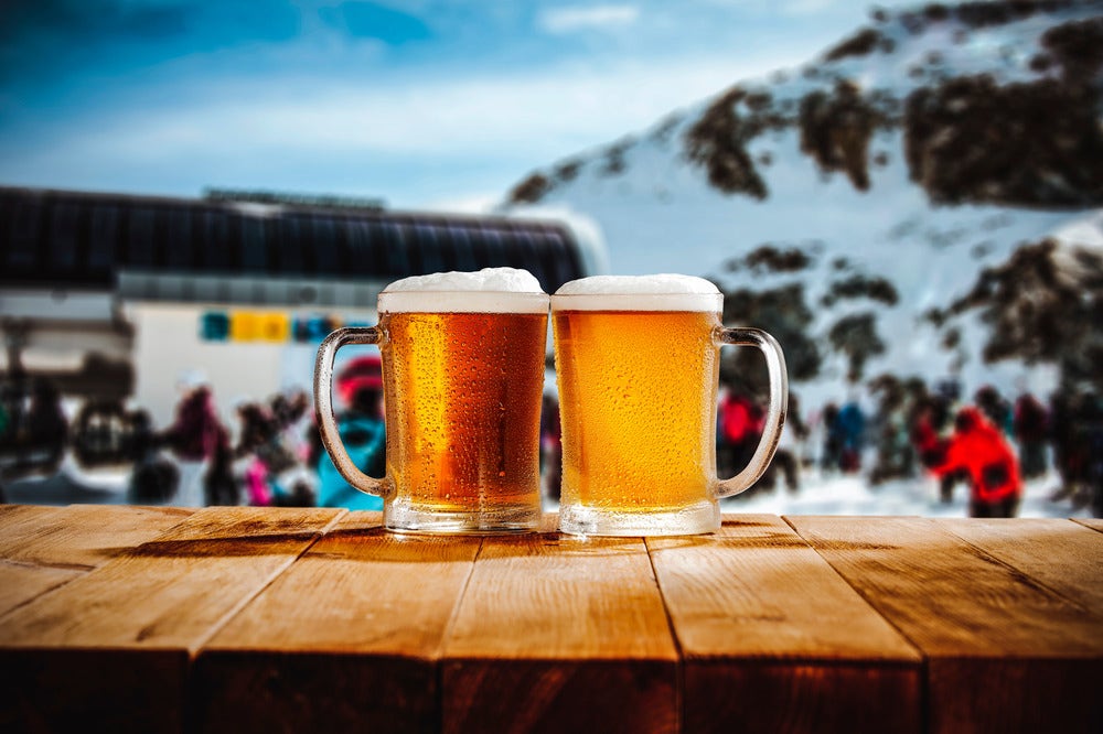 Two frosty mugs of golden beer on a wooden table with a snowy mountain backdrop and people enjoying a winter scene in the background.