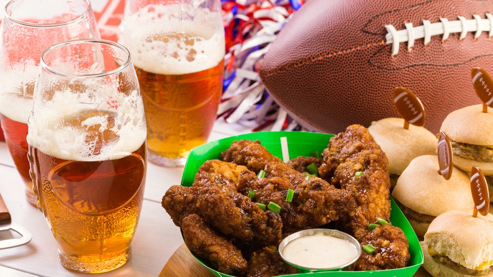 Glasses of beer with crispy buffalo wings and sliders on a game-day table, decorated with football-themed party elements.