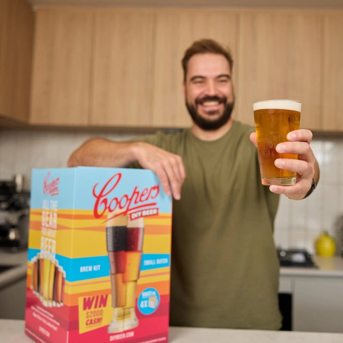 Man holding a Coopers beer box and a glass of beer in a kitchen.