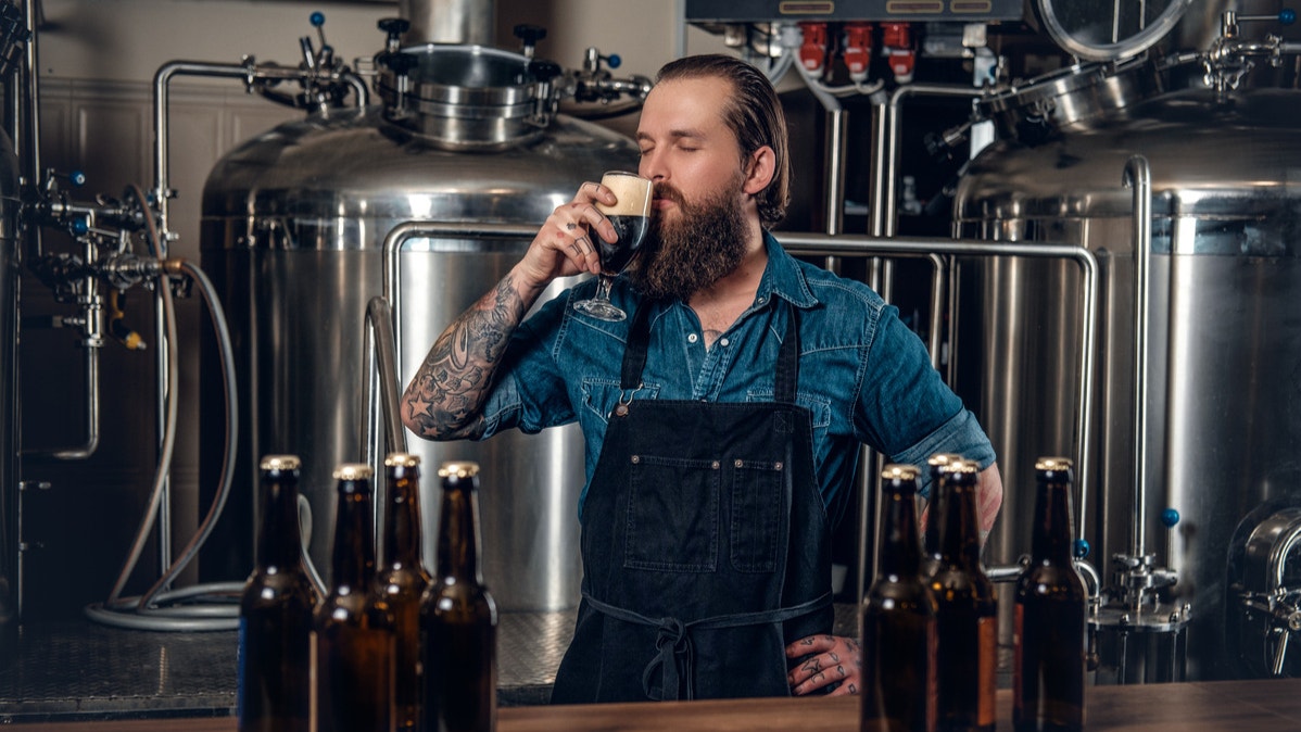 Bearded homebrewer in a denim shirt and apron tasting a dark beer in a brewery, surrounded by beer bottles and stainless steel brewing equipment.