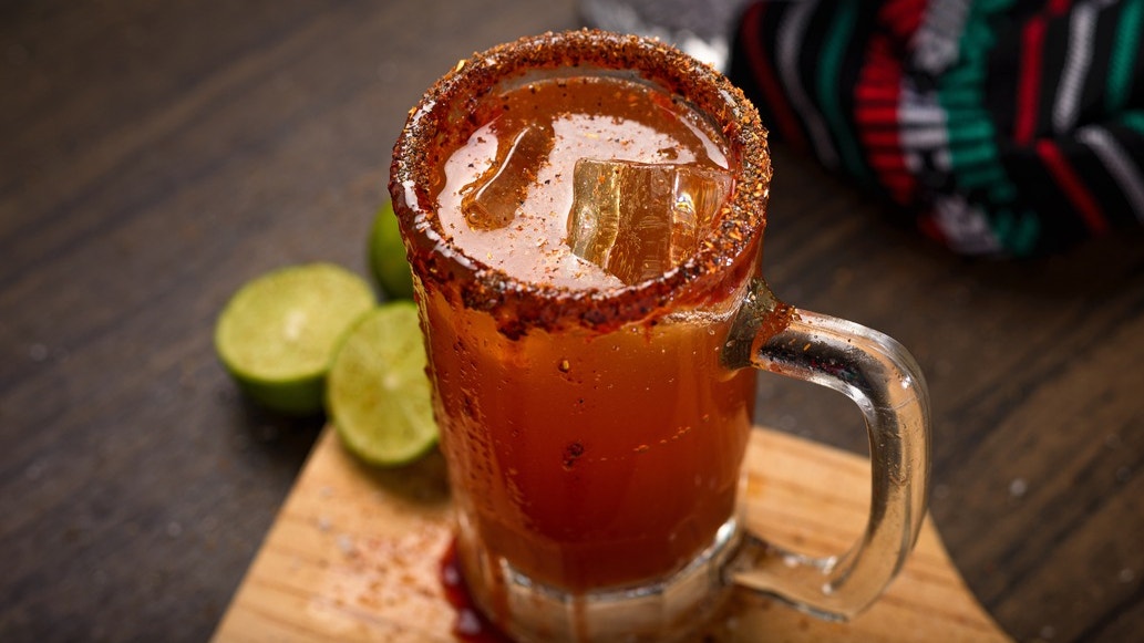 Michelada beer cocktail served in a glass mug with Tajín-rim, ice cubes, and fresh lime on a wooden board